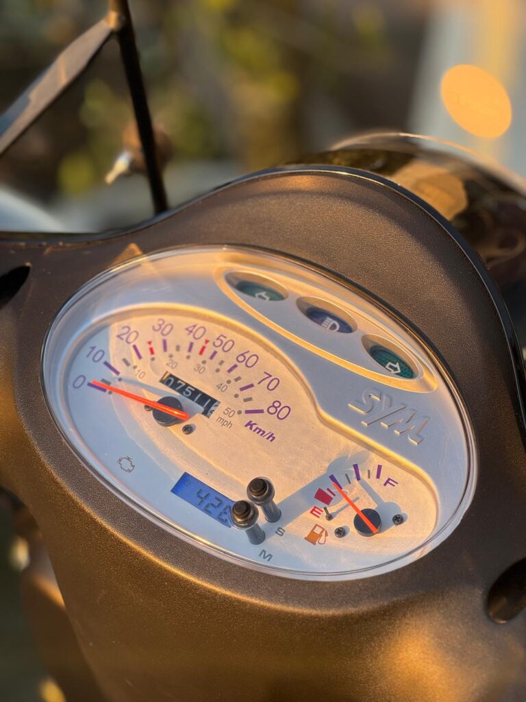 Close-up van het SYM-scooter dashboard met snelheidsmeter, brandstofmeter en km-teller in zonlicht.