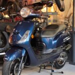 Blauwe retro-scooter op de middenbok in een showroom met andere scooters op de achtergrond.