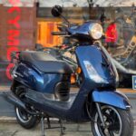 Donkerblauwe scooter op de middenbok voor een winkelruit met Kymco-logo, gezien vanaf rechtsvoor.