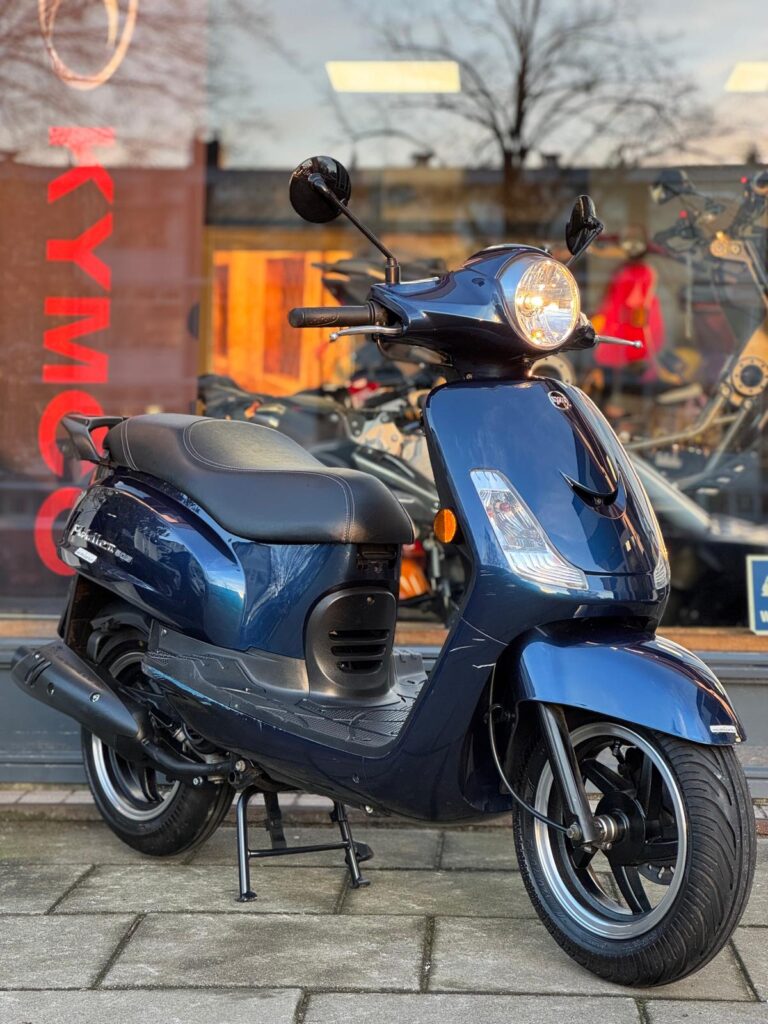 Donkerblauwe scooter op de middenbok voor een winkelruit met Kymco-logo, gezien vanaf rechtsvoor.
