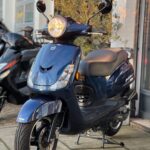 Donkerblauwe scooter met koplamp aan, op standaard voor een winkel.