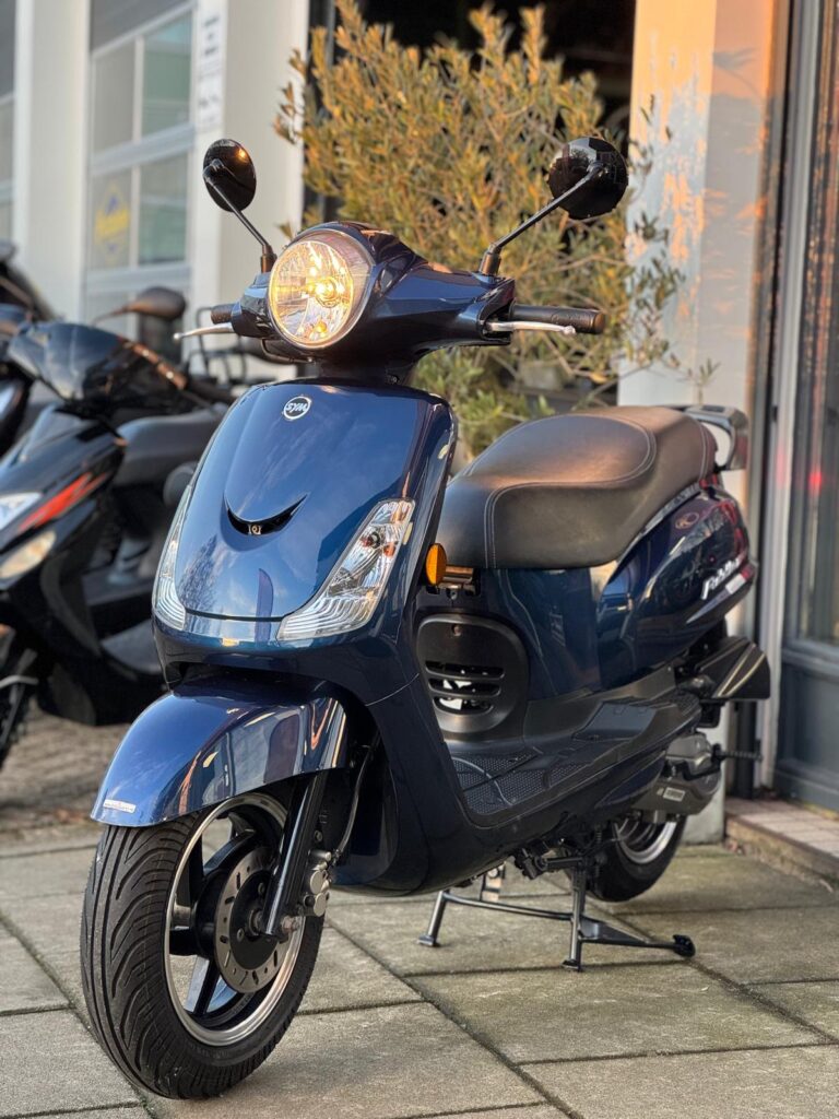 Donkerblauwe scooter met koplamp aan, op standaard voor een winkel.
