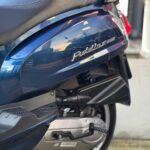 Close-up van de zijkant van een donkerblauwe scooter met zadel, uitlaat, motorblok en achterwiel buiten.