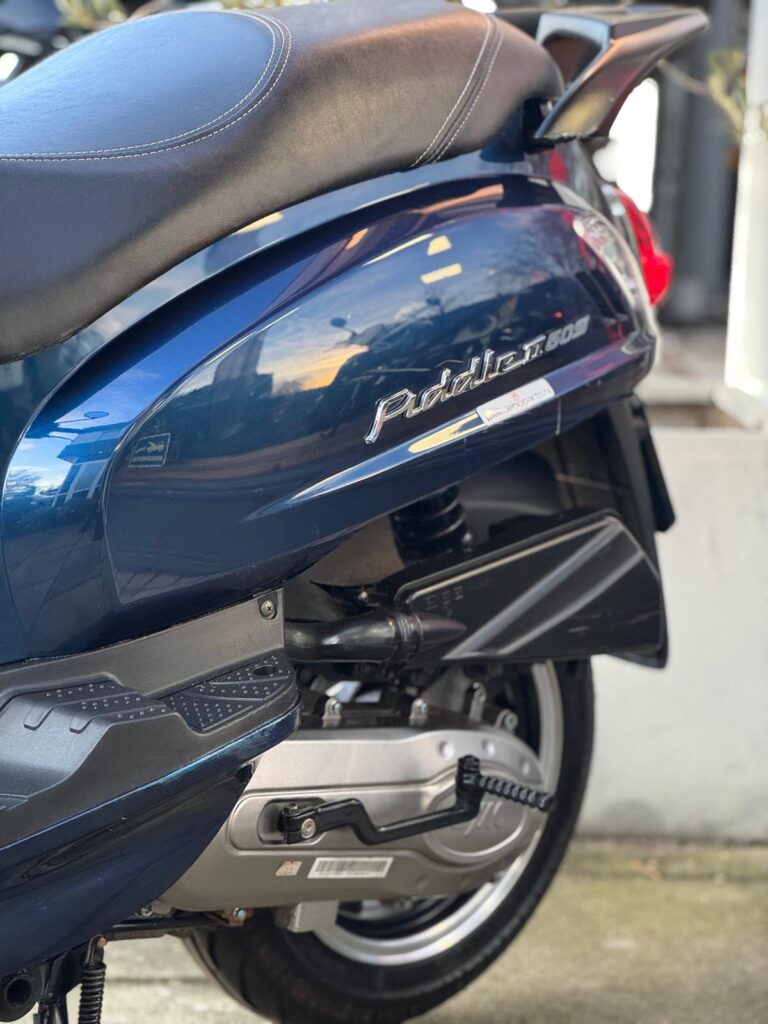 Close-up van de zijkant van een donkerblauwe scooter met zadel, uitlaat, motorblok en achterwiel buiten.
