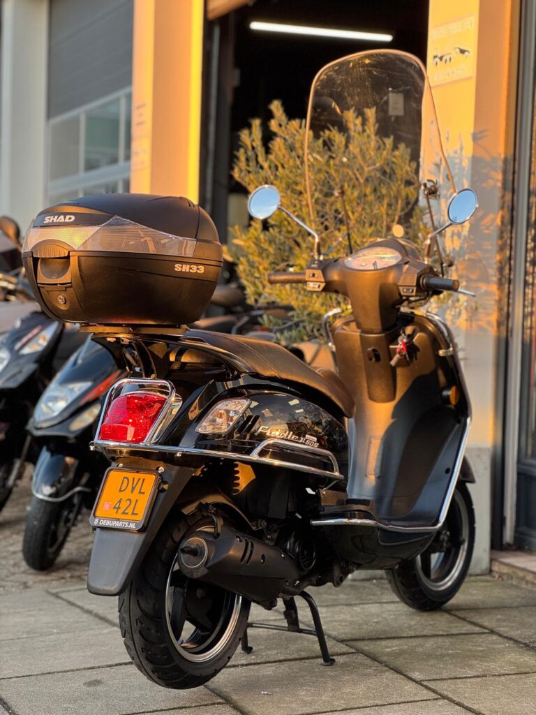 Zwarte scooter met topkoffer en hoog windscherm, geparkeerd voor een werkplaats in avondzon.