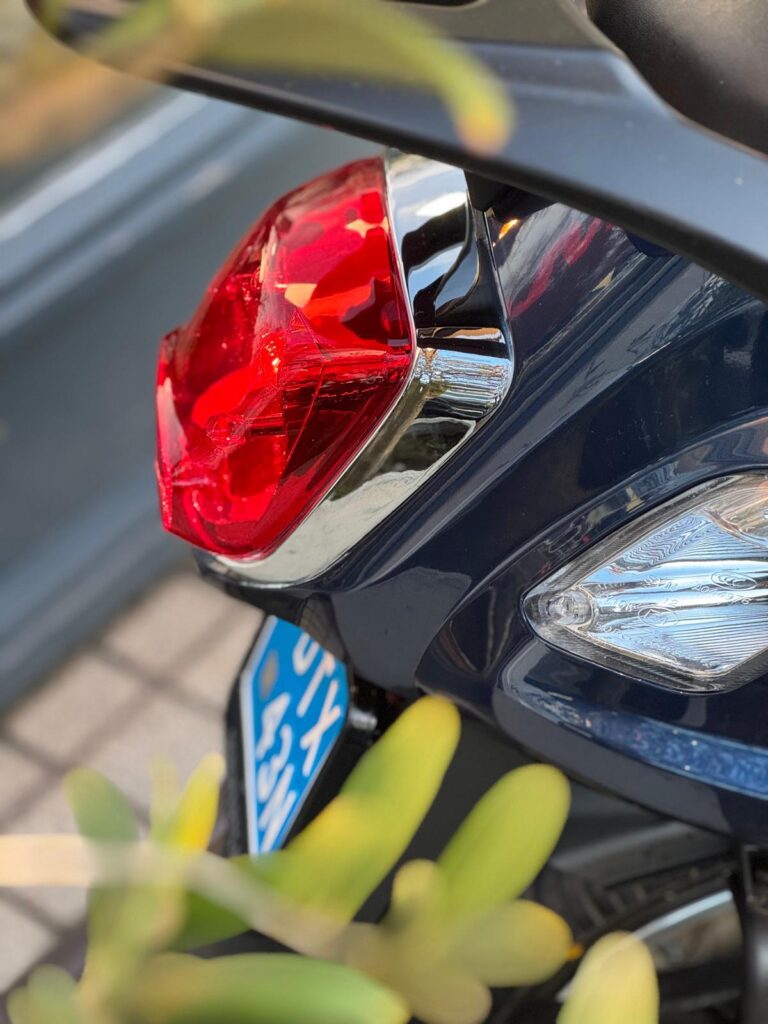 Close-up van het rode achterlicht met chroomrand en deel van kenteken van een blauwe scooter.