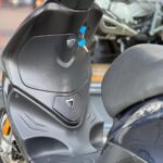 Close-up van zwarte scooter met blauwe sleutel in het contact, met zicht op voetplaat en zadel.