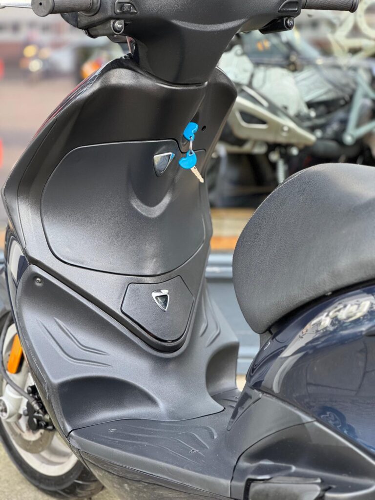 Close-up van zwarte scooter met blauwe sleutel in het contact, met zicht op voetplaat en zadel.