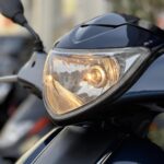 Close-up van een brandende koplamp van een donkerblauwe scooter, met spiegel op de achtergrond.