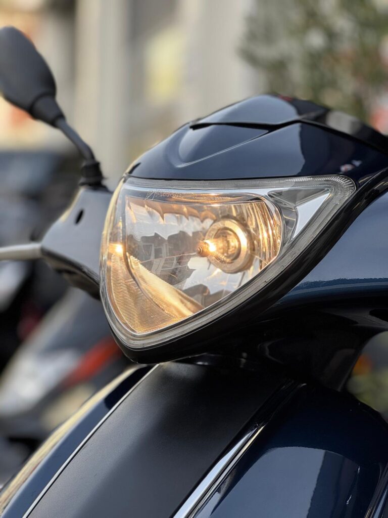 Close-up van een brandende koplamp van een donkerblauwe scooter, met spiegel op de achtergrond.