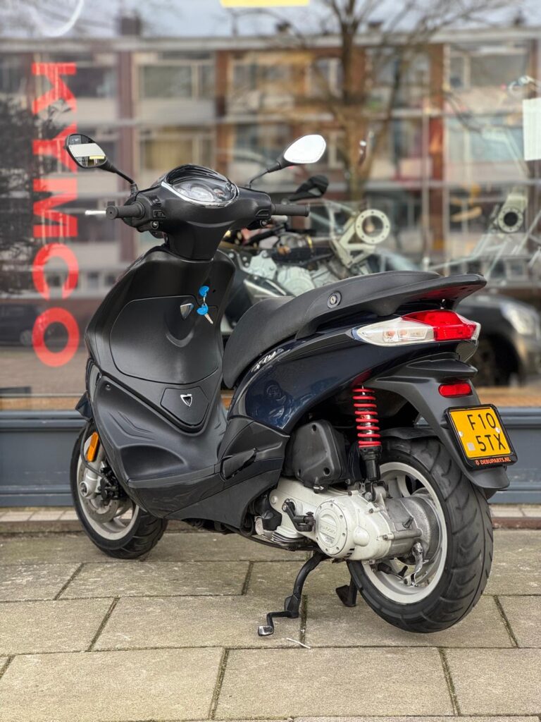 Zwarte scooter op zijn standaard, geparkeerd op het trottoir voor een winkelraam met Kymco-logo.