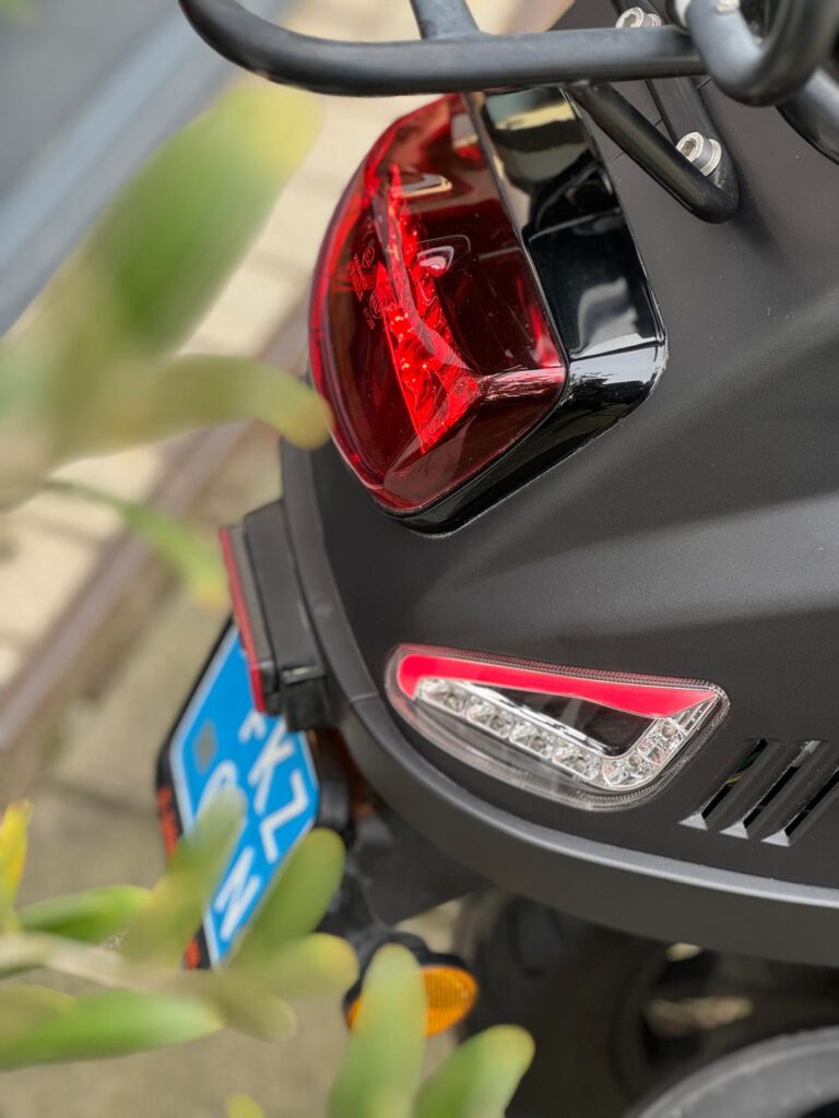 Close-up van de achterkant van een zwarte scooter met rood achterlicht, richtingaanwijzer en blauwe kentekenplaat.