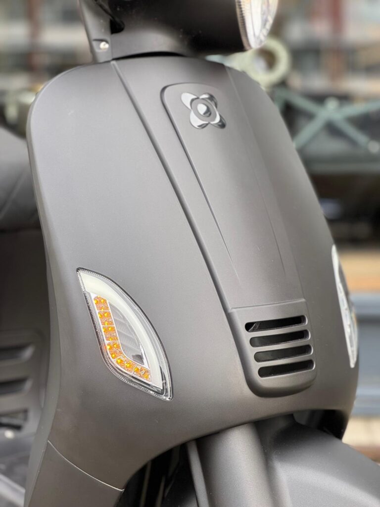 Close-up van de matte zwarte voorkant van een scooter met LED knipperlichten en ventilatierooster.