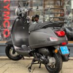 Mat zwarte AGM scooter met bagagedrager en NL kenteken, geparkeerd voor een winkel.