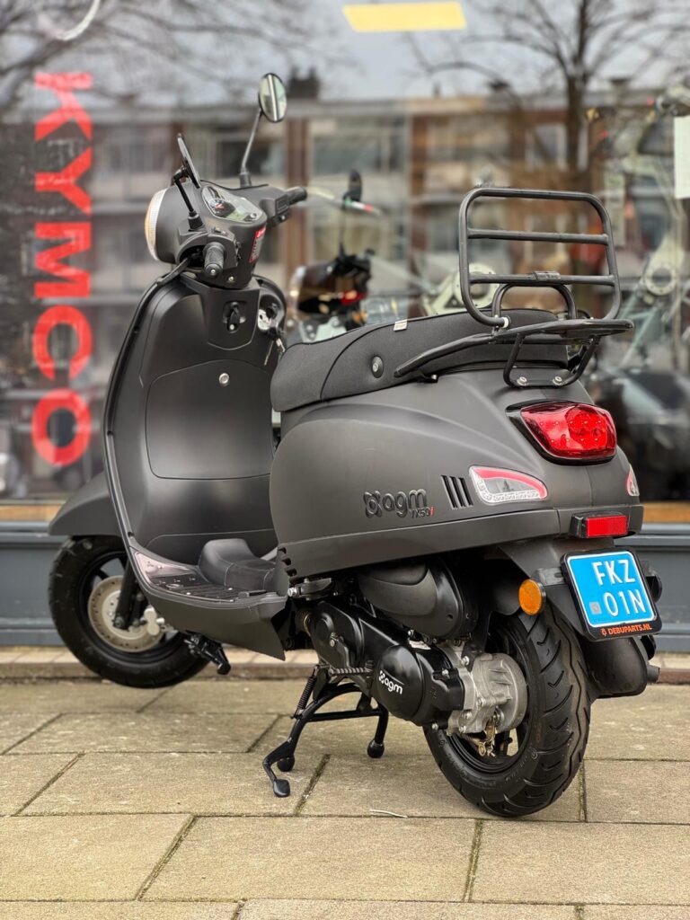 Mat zwarte AGM scooter met bagagedrager en NL kenteken, geparkeerd voor een winkel.
