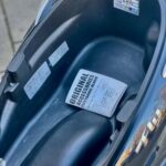 Open opbergvak onder het zadel van een scooter met handleiding en service-sticker.