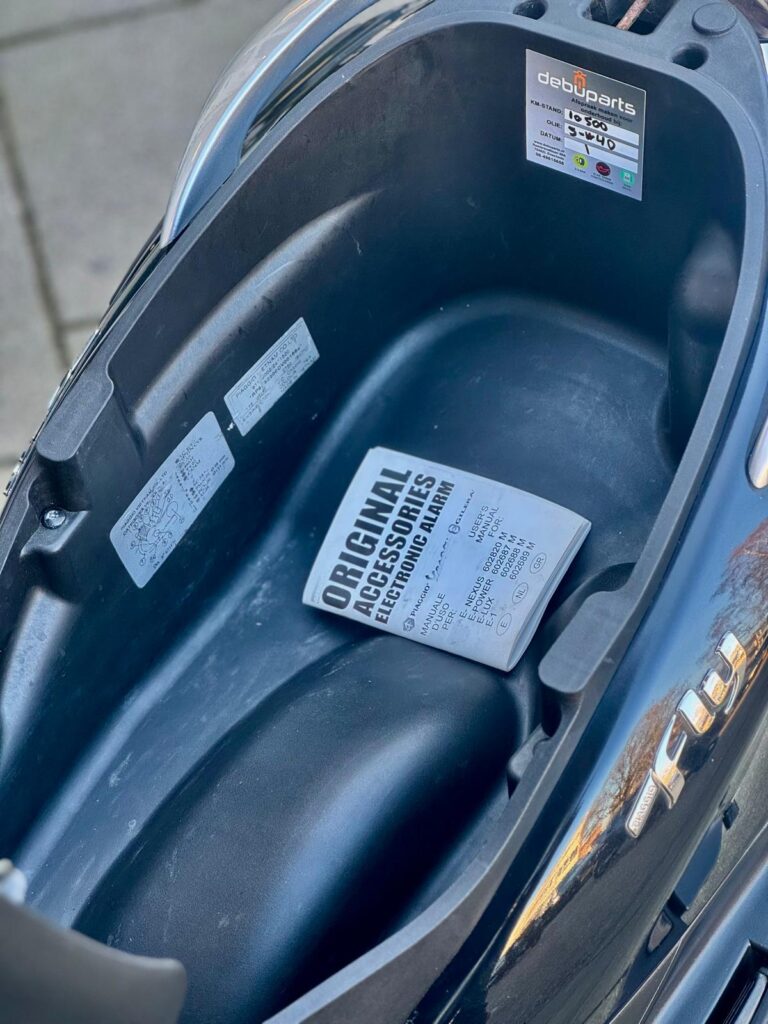 Open opbergvak onder het zadel van een scooter met handleiding en service-sticker.