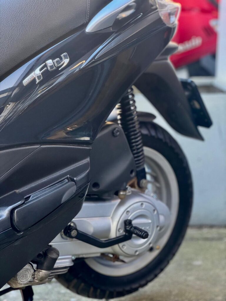 Close-up van de achterkant en motor van een zwarte scooter met achterwiel, vering en kickstarter.