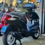 Zwarte scooter met blauw NL-kenteken, geparkeerd op stoep voor winkel, andere scooters op achtergrond.