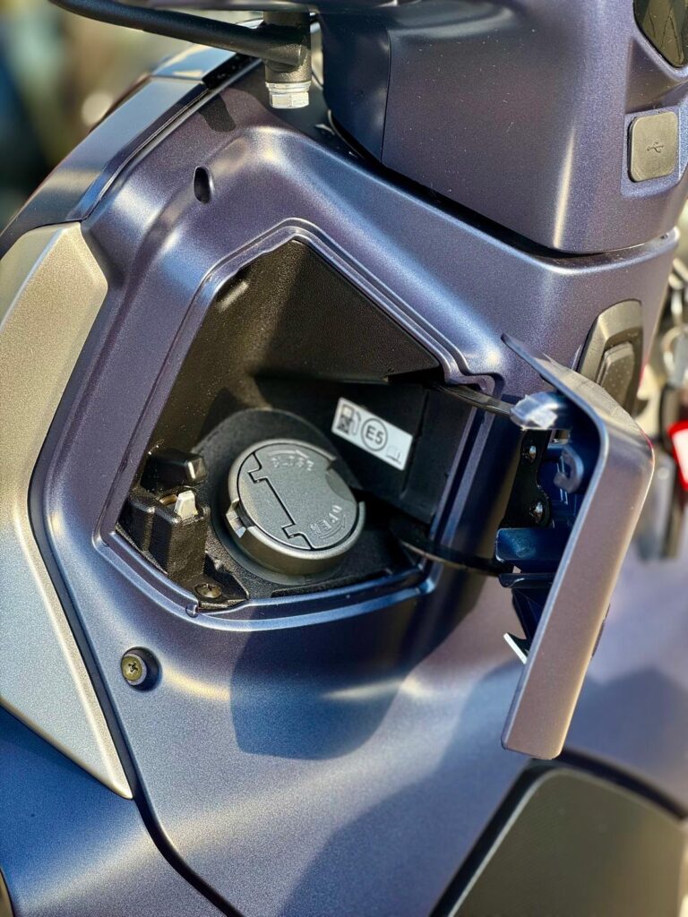 Close-up van het geopende brandstofklepje van een scooter, met dop in een klein opbergvak.