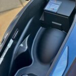 Open opbergvak onder het zadel van een scooter met zwarte accubox en stickers.
