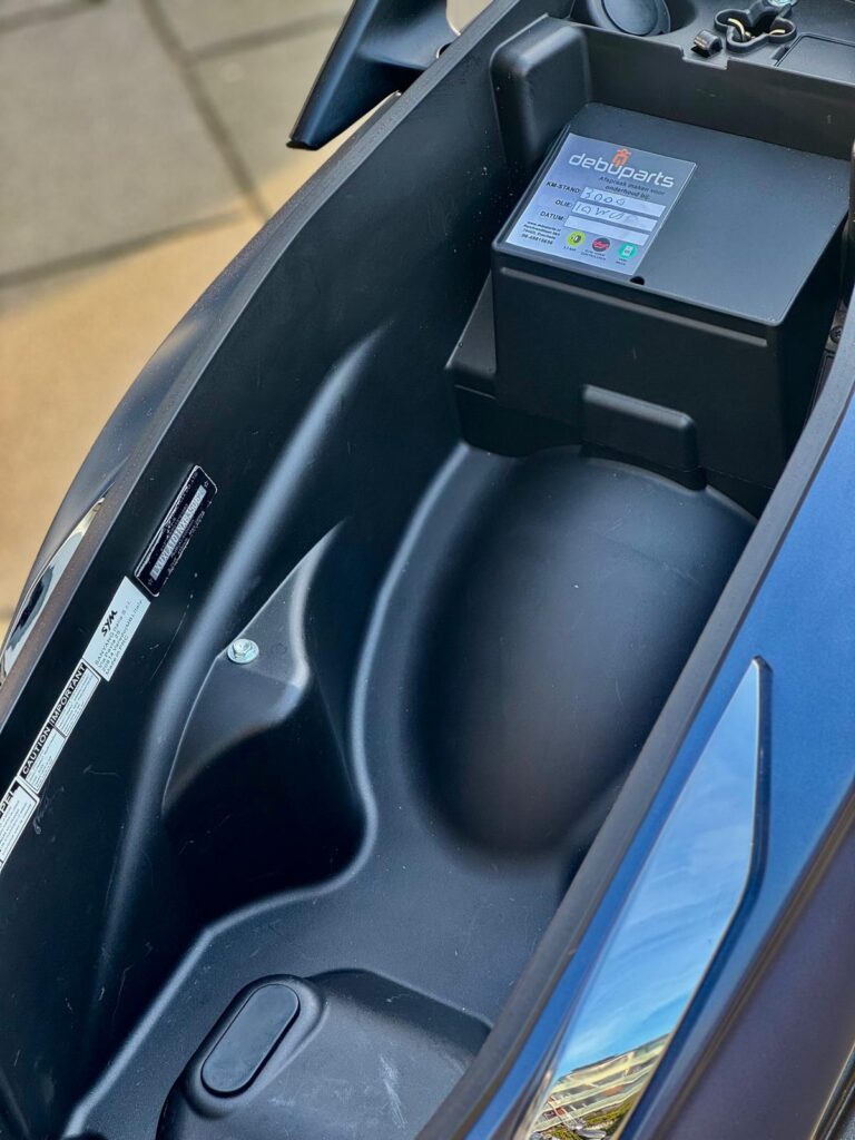 Open opbergvak onder het zadel van een scooter met zwarte accubox en stickers.