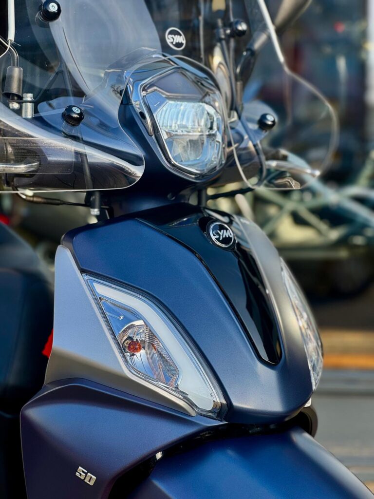 Close-up van een blauwe SYM-scooter met windscherm en koplamp, vooraanzicht.