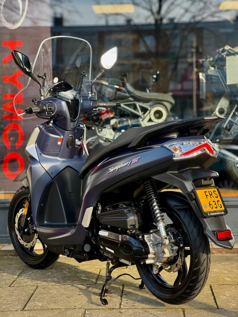 Donkergrijze Kymco-scooter met windscherm, op de standaard voor een showroom met motoren.