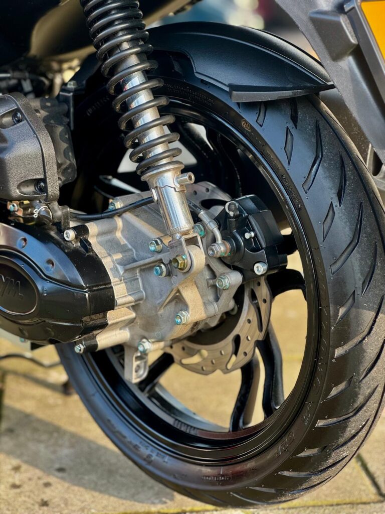 Close-up van het achterwiel van een scooter met schokdemper, remschijf en brede band.