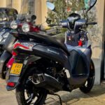 Zwarte scooter met ruit en spiegels geparkeerd buiten een winkel, met andere scooters op de achtergrond.