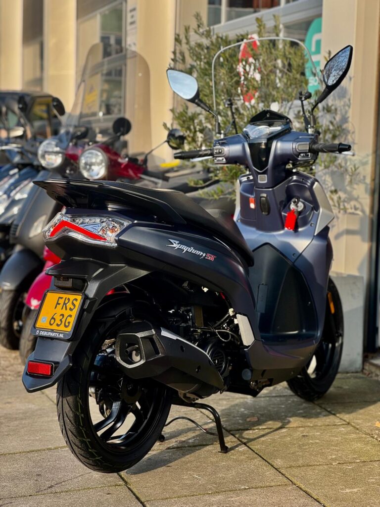 Zwarte scooter met ruit en spiegels geparkeerd buiten een winkel, met andere scooters op de achtergrond.