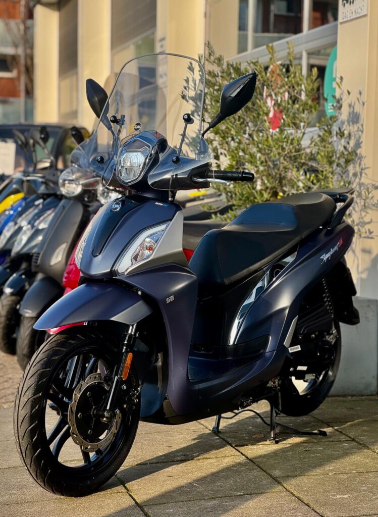 Donkerblauwe scooter met hoog windscherm, geparkeerd buiten bij een rij andere scooters.