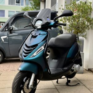 Groenblauwe scooter met ruitje en schijfrem, geparkeerd op het trottoir bij een appartementencomplex.