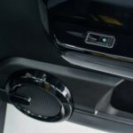 Detail van een autodeurpaneel met verlichte elektrische raamknop en ronde luidspreker.