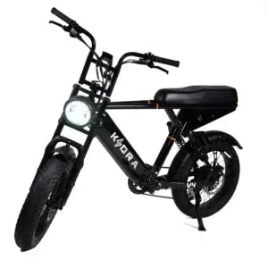 Zwarte mopedstijl e-bike met dikke banden, brandende ronde koplamp en lang zadel op witte achtergrond.