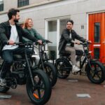 Drie mensen fietsen lachend op zwarte elektrische fatbikes door een woonstraat.