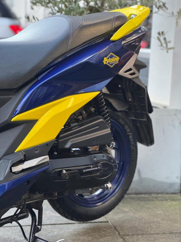 Close-up van blauwe en gele scooter met Putoline-sticker, achterwiel en zadel zichtbaar.