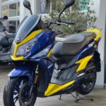 Blauw-gele sportieve scooter met dubbele koplamp, geparkeerd op de stoep bij een showroom.