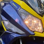 Close-up van de voorkant van een blauw-gele SYM scooter met brandende koplamp.