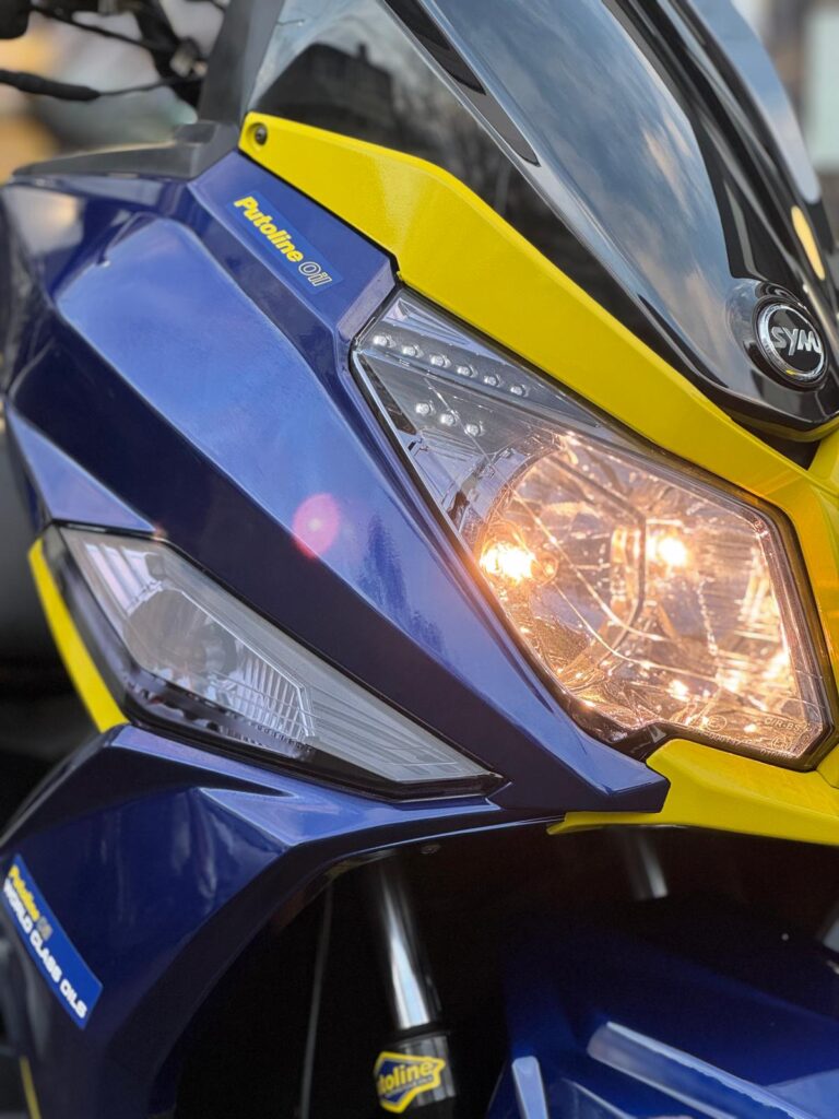 Close-up van de voorkant van een blauw-gele SYM scooter met brandende koplamp.