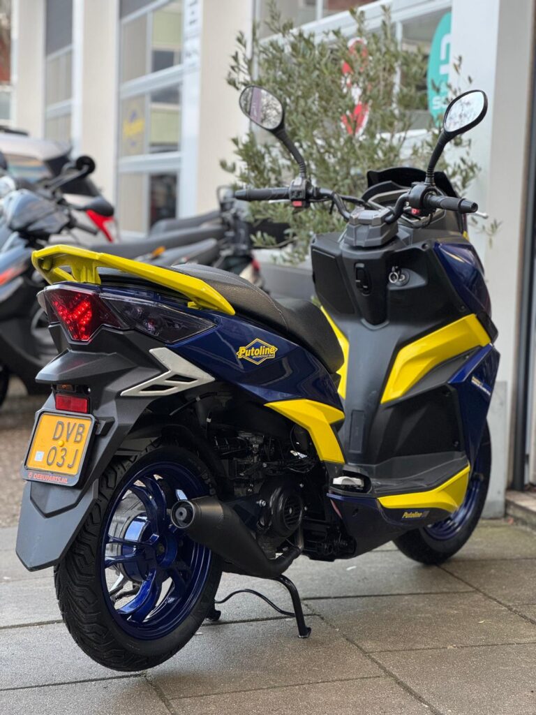 Blauw-gele scooter met Putoline-styling, geparkeerd op de stoep bij een scootershop.