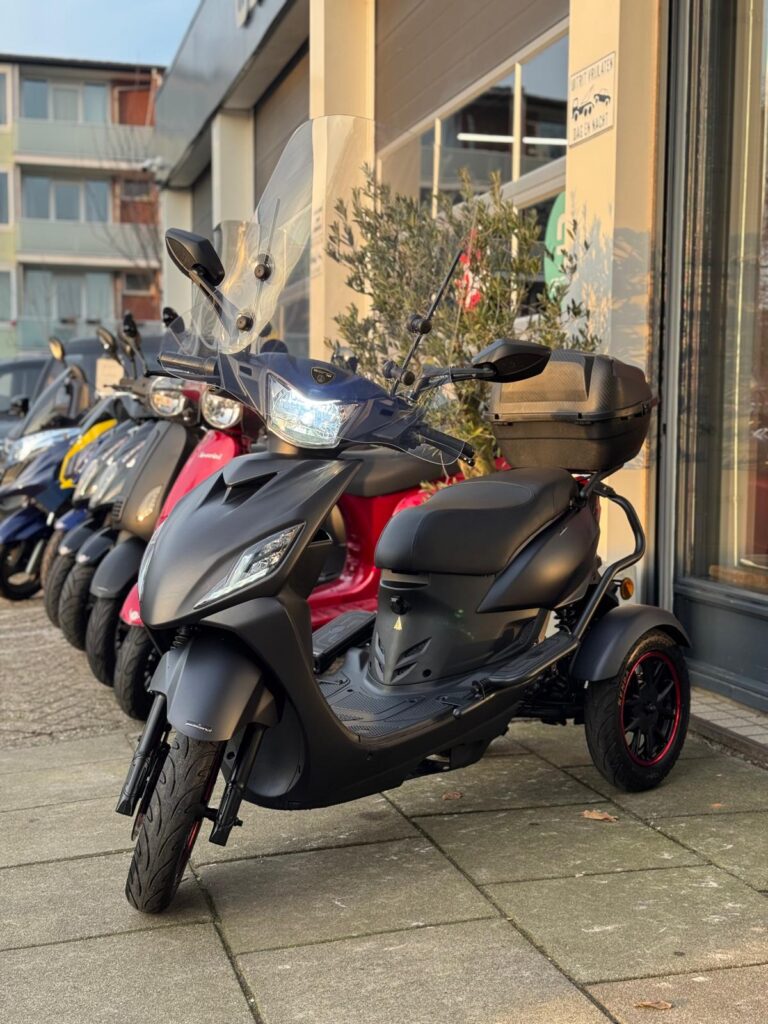 Matgrijze driewielscooter met windscherm en topkoffer, geparkeerd bij een scooterzaak.