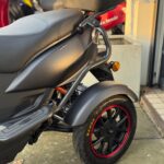 Detail van een matzwarte scooter met topkoffer, voorwiel met rode rand en Kenda-band, geparkeerd buiten.