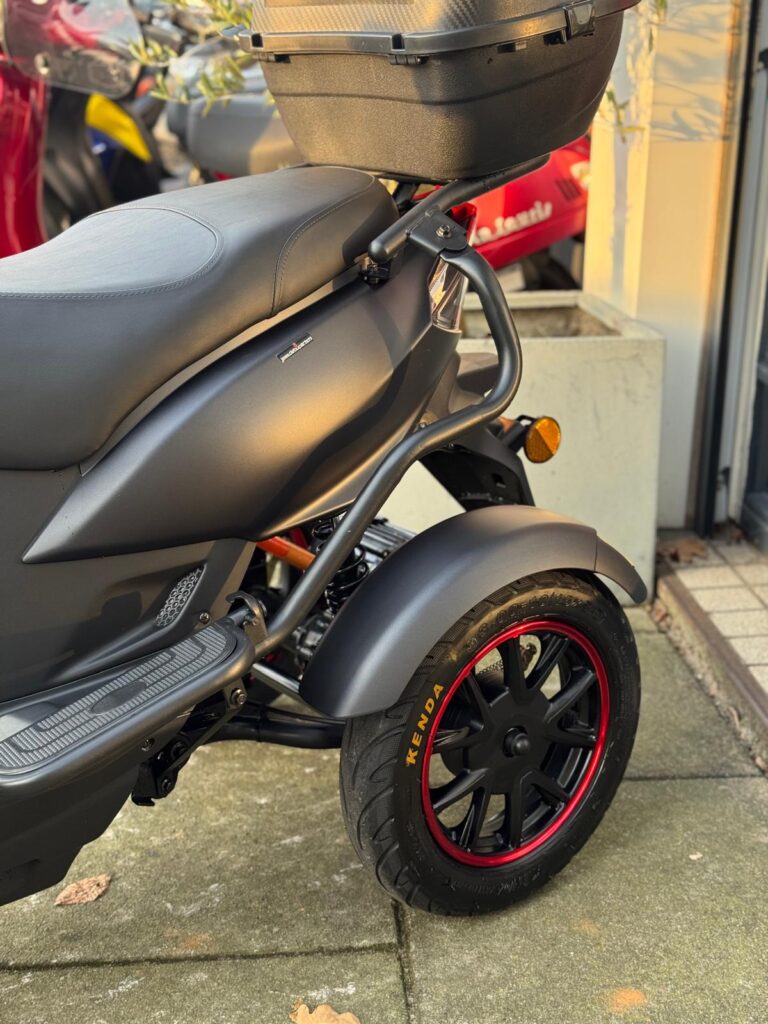 Detail van een matzwarte scooter met topkoffer, voorwiel met rode rand en Kenda-band, geparkeerd buiten.