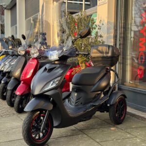 Mat zwarte driewielige scooter met windscherm en topkoffer, geparkeerd voor een scootershop.