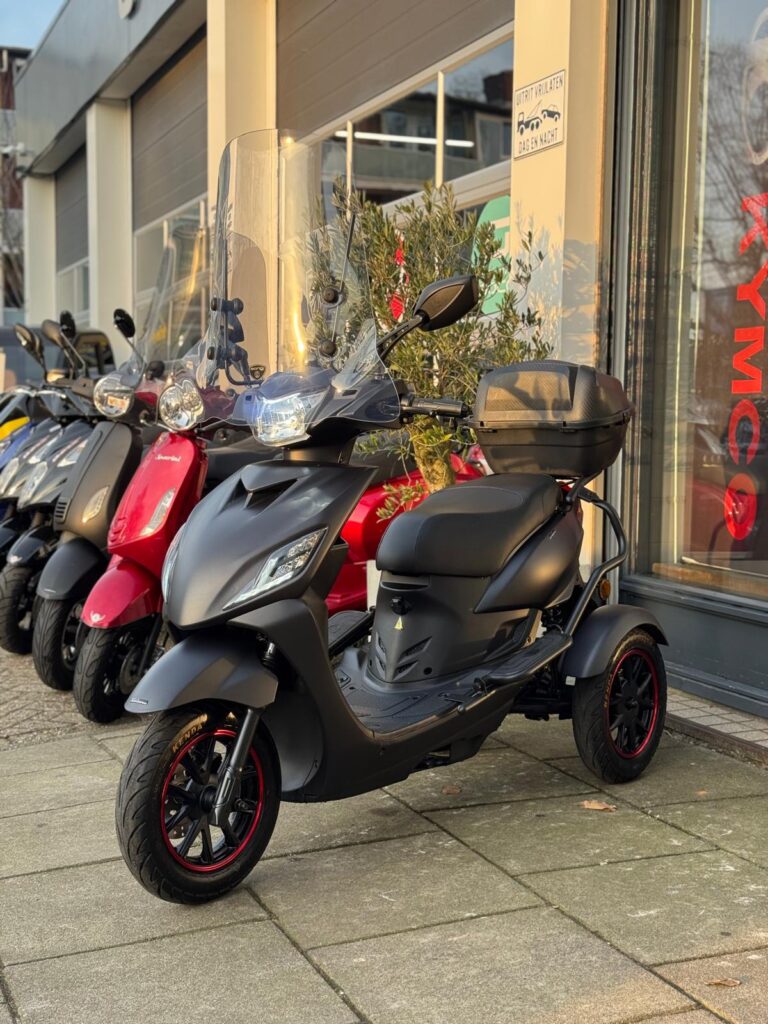 Mat zwarte driewielige scooter met windscherm en topkoffer, geparkeerd voor een scootershop.