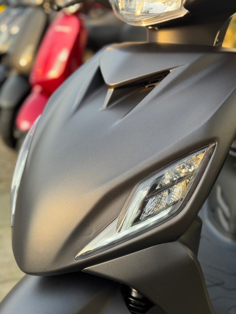 Close-up van de voorkant van een matte zwarte scooter met LED-koplampen en onscherpe scooters op de achtergrond.