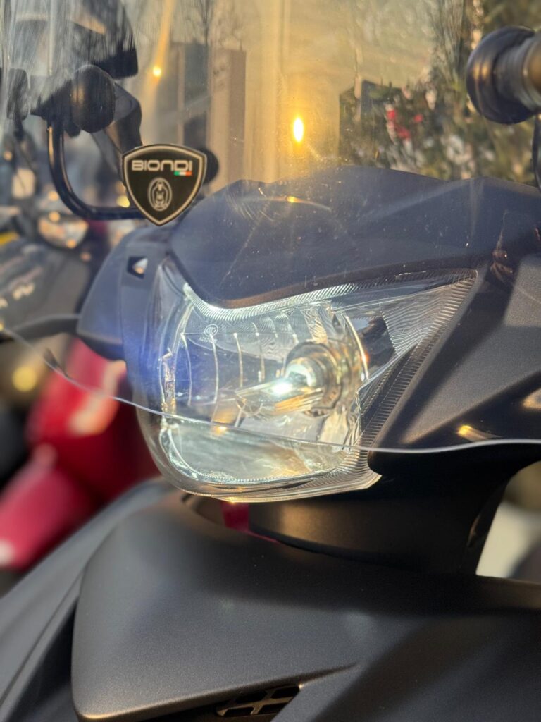 Close-up van scooterkoplamp met windscherm en Biondi-sticker in warm zonlicht