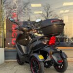 Zwarte driewielige scooter met topkoffer, geparkeerd voor een winkelpui met grote ramen.