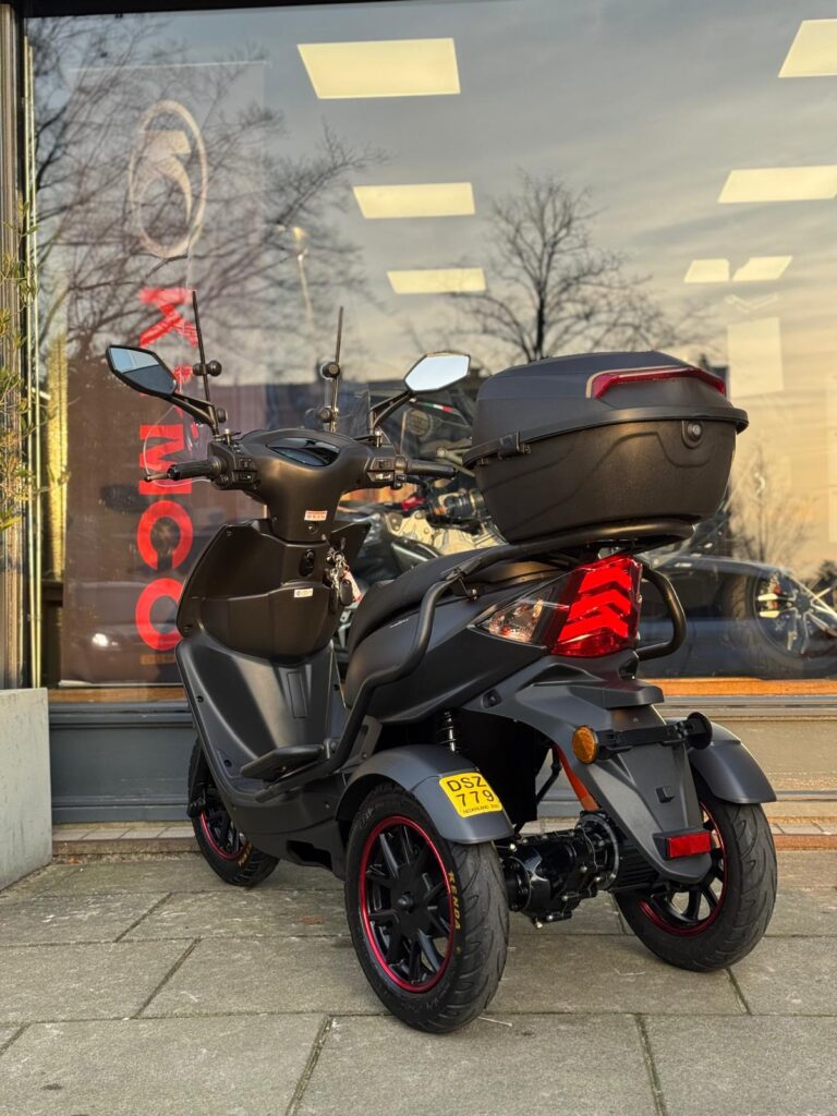 Zwarte driewielige scooter met topkoffer, geparkeerd voor een winkelpui met grote ramen.